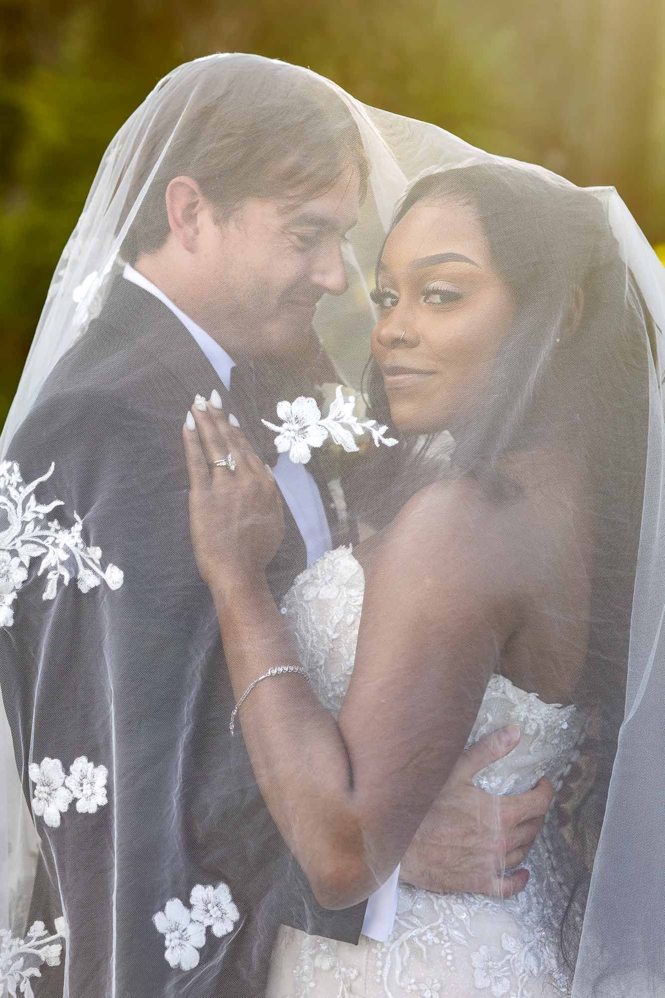 Interracial couple sharing a romantic moment under a floral lace wedding veil outdoors in golden sunlight.