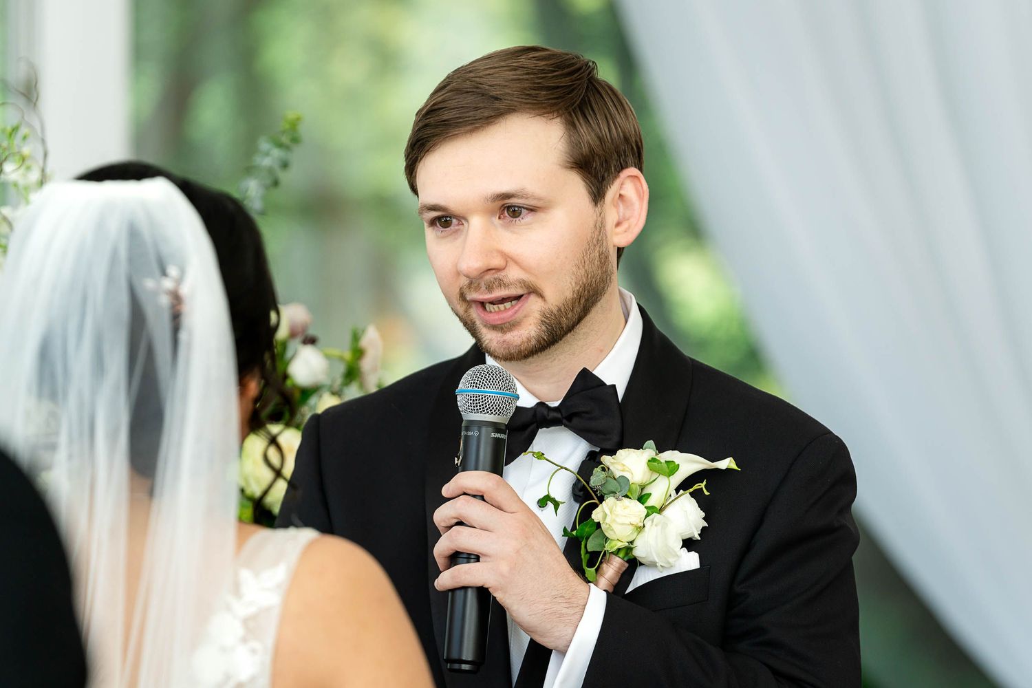 Groom in black tuxedo speaking into microphone at wedding ceremony with boutonniere and bride in veil.