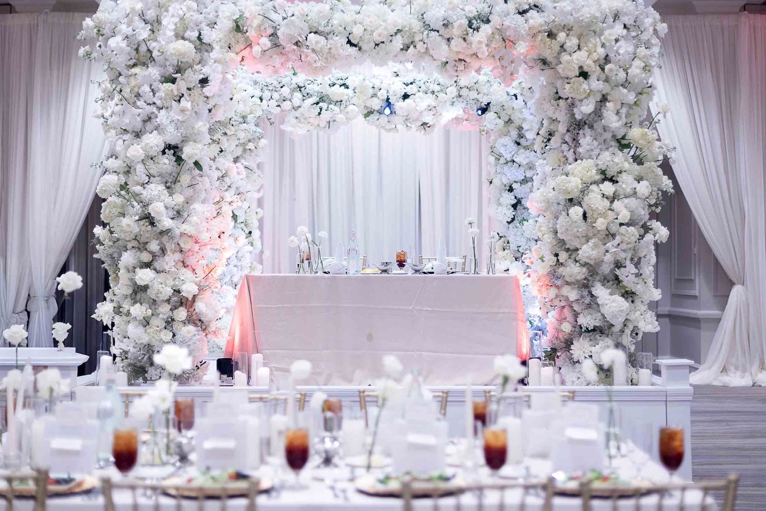 Elegant white wedding reception with floral arch, draped ceiling, and beautifully set tables adorned with white flowers.
