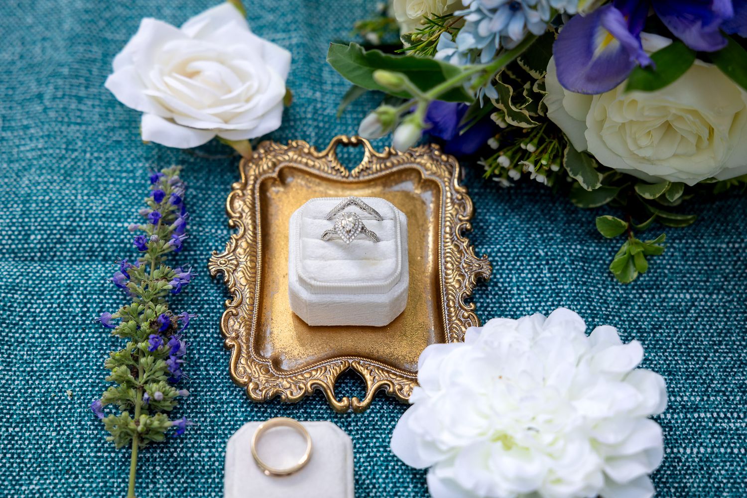 Diamond engagement ring in ornate gold frame box surrounded by white roses, lavender, and blue flowers on teal background.