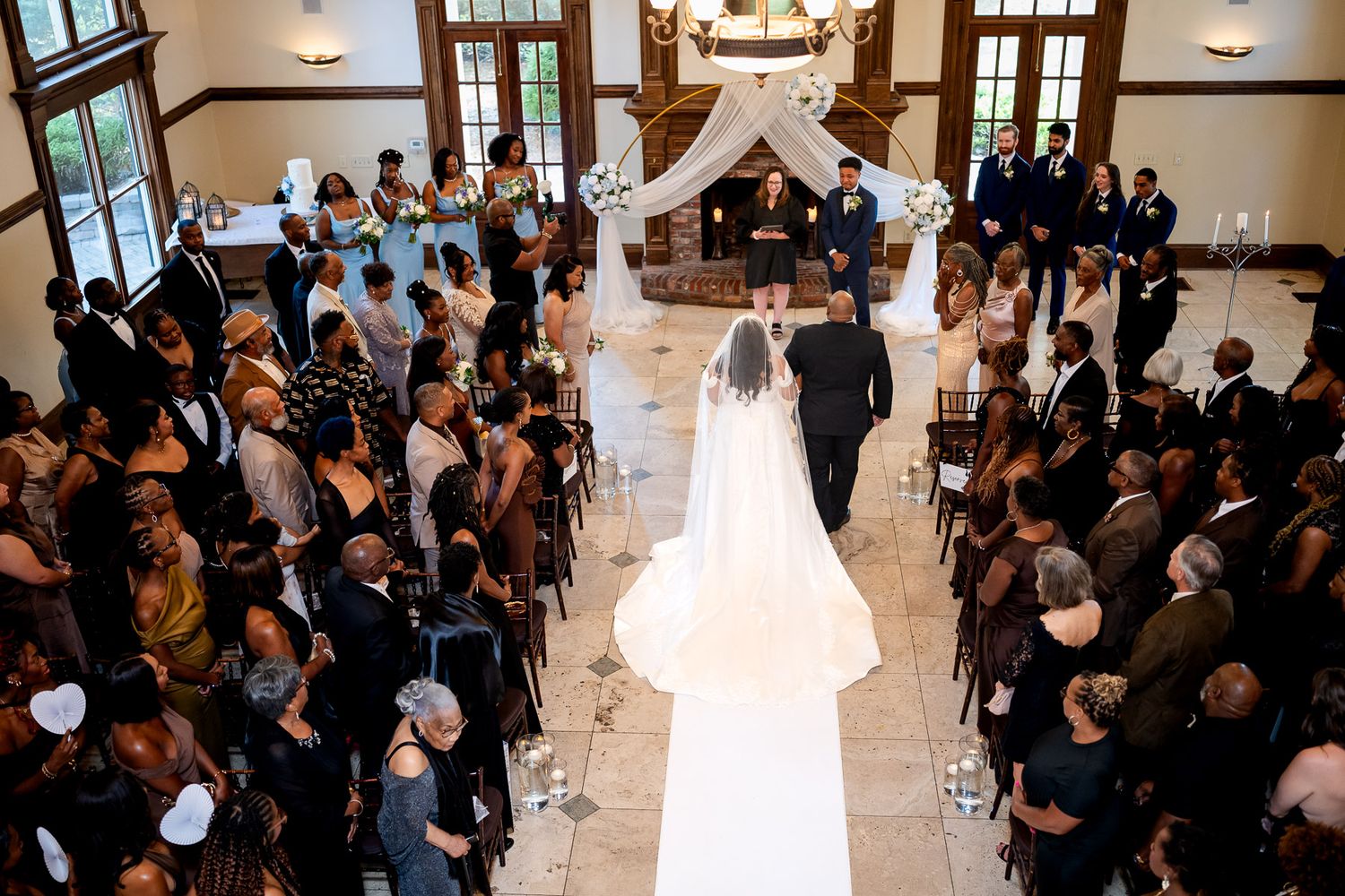 Elegant wedding ceremony with bride and groom walking down white aisle in grand venue with floral arch.