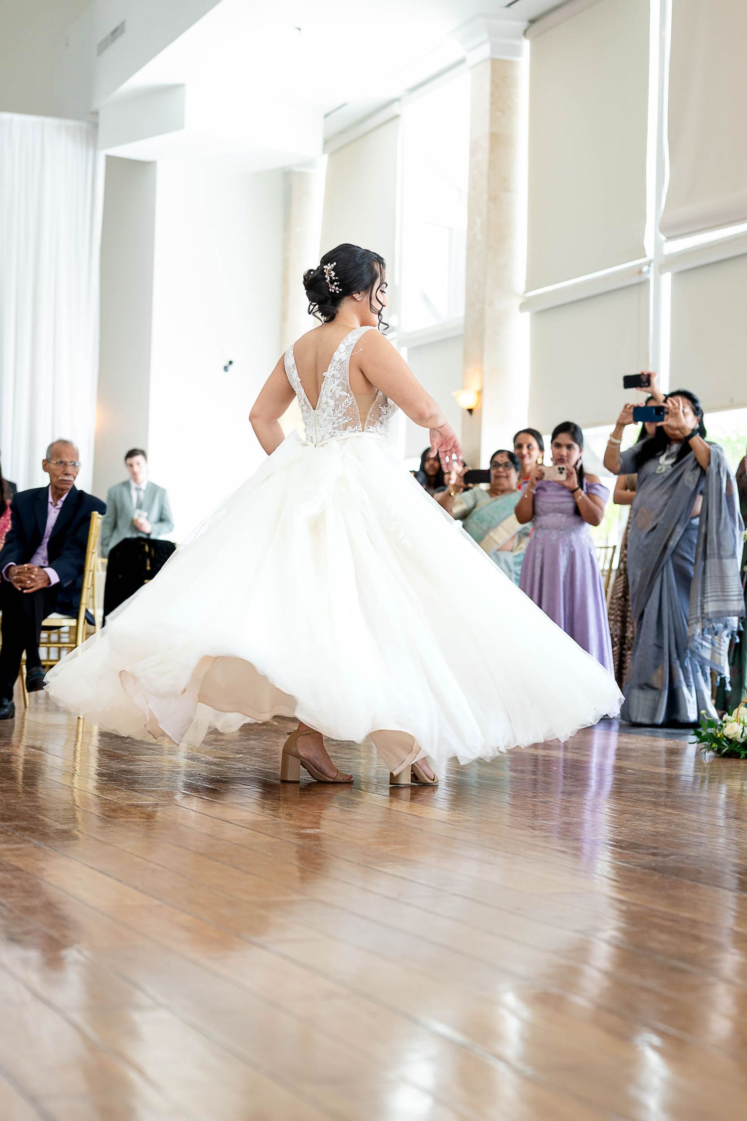 Bride twirls in a stunning white ball gown during first dance as wedding guests watch and photograph the moment.
