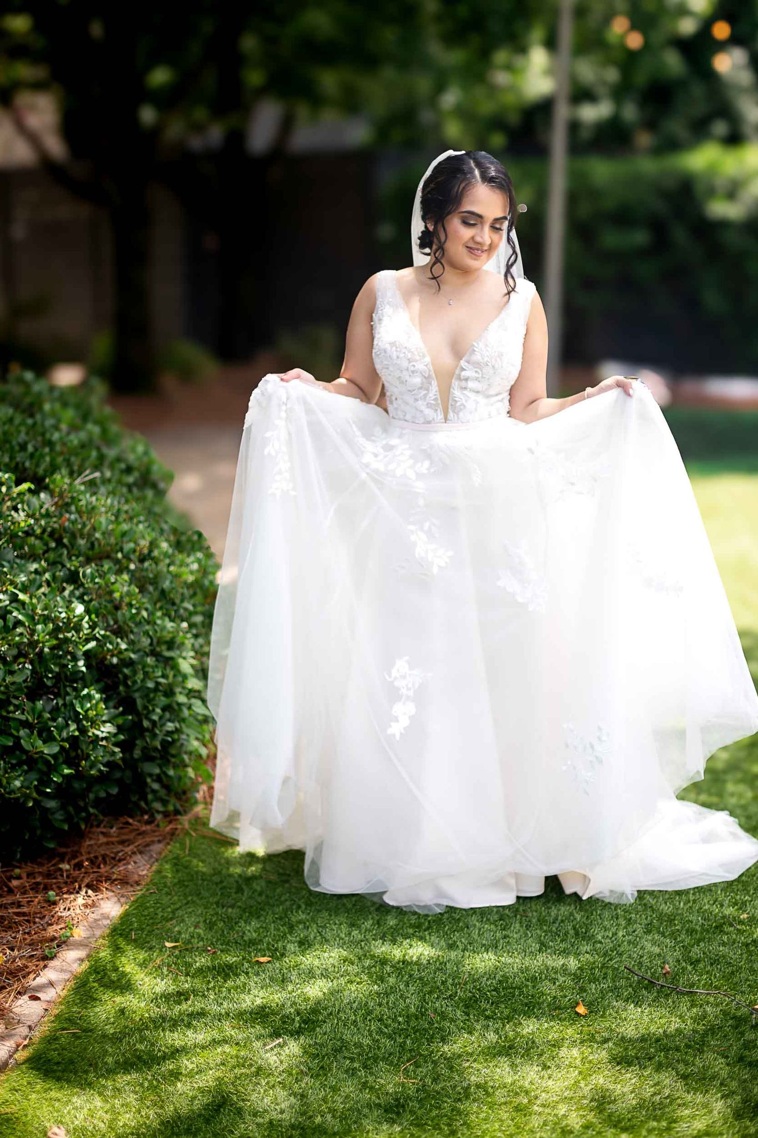 Bride in white lace deep V-neck ballgown lifting skirt while standing on green lawn.