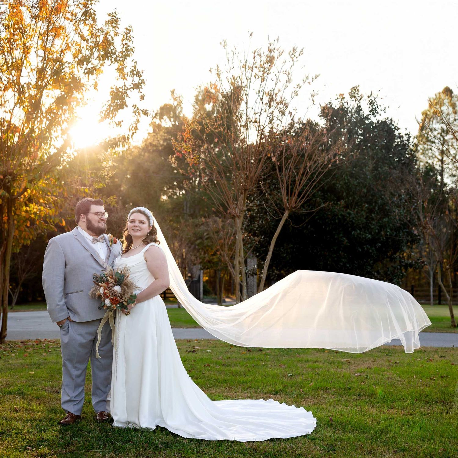 Bride and groom pose at golden hour on green lawn, her veil flowing in wind, surrounded by autumn trees.