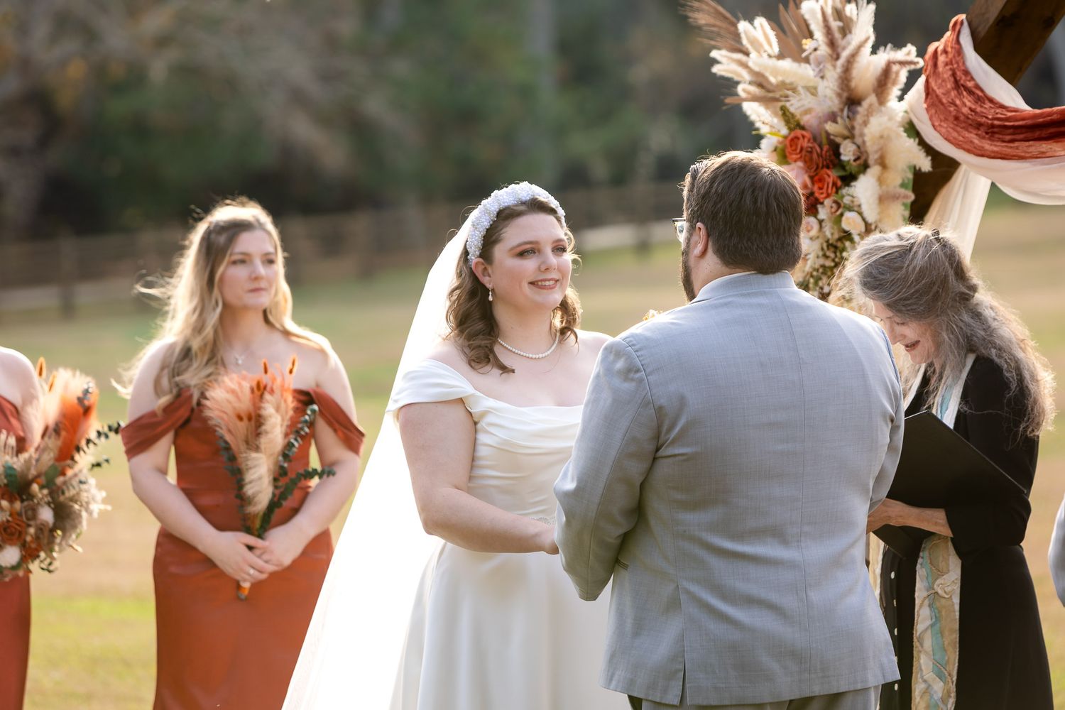 Bride and groom exchange vows outdoors with bridesmaid in rust dress and officiant during fall wedding ceremony.