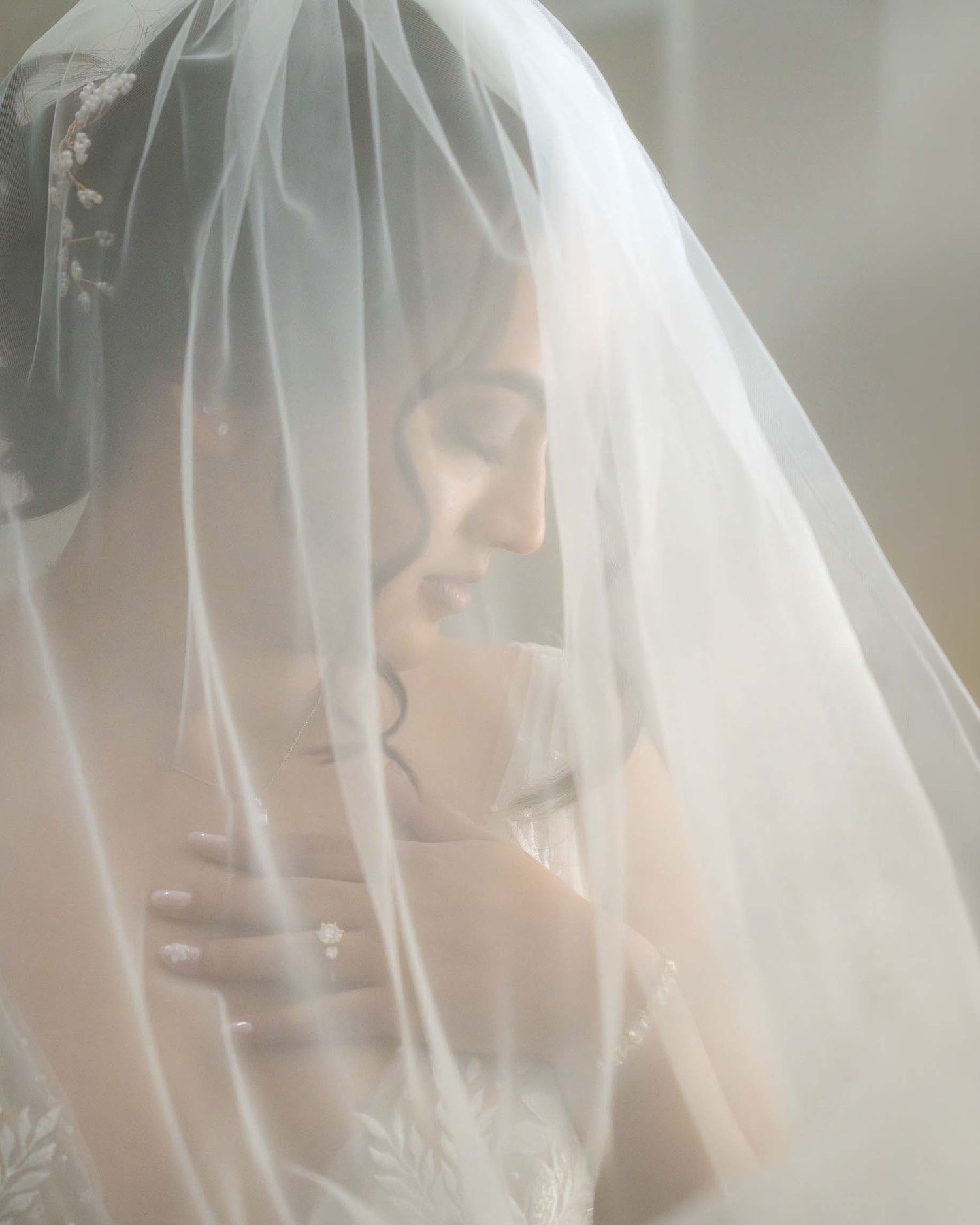 A bride draped in a sheer white veil, eyes closed, hand on chest, in a soft dreamy bridal portrait.