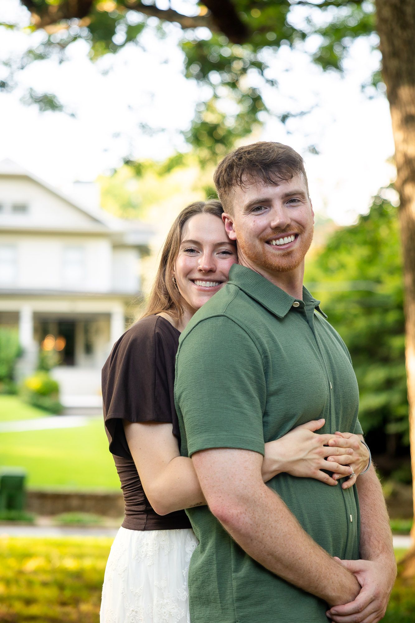 A smiling couple embraces outdoors near a house, woman in brown top hugs man in green polo shirt.