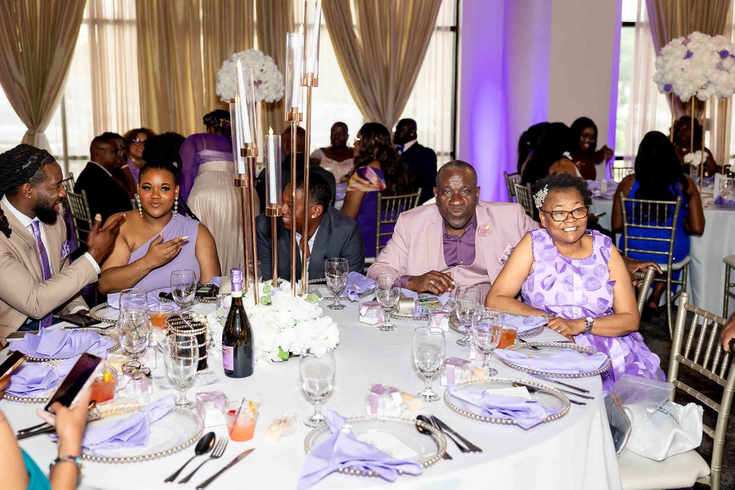 Wedding guests seated at lavender-themed reception tables with tall floral centerpieces and champagne.