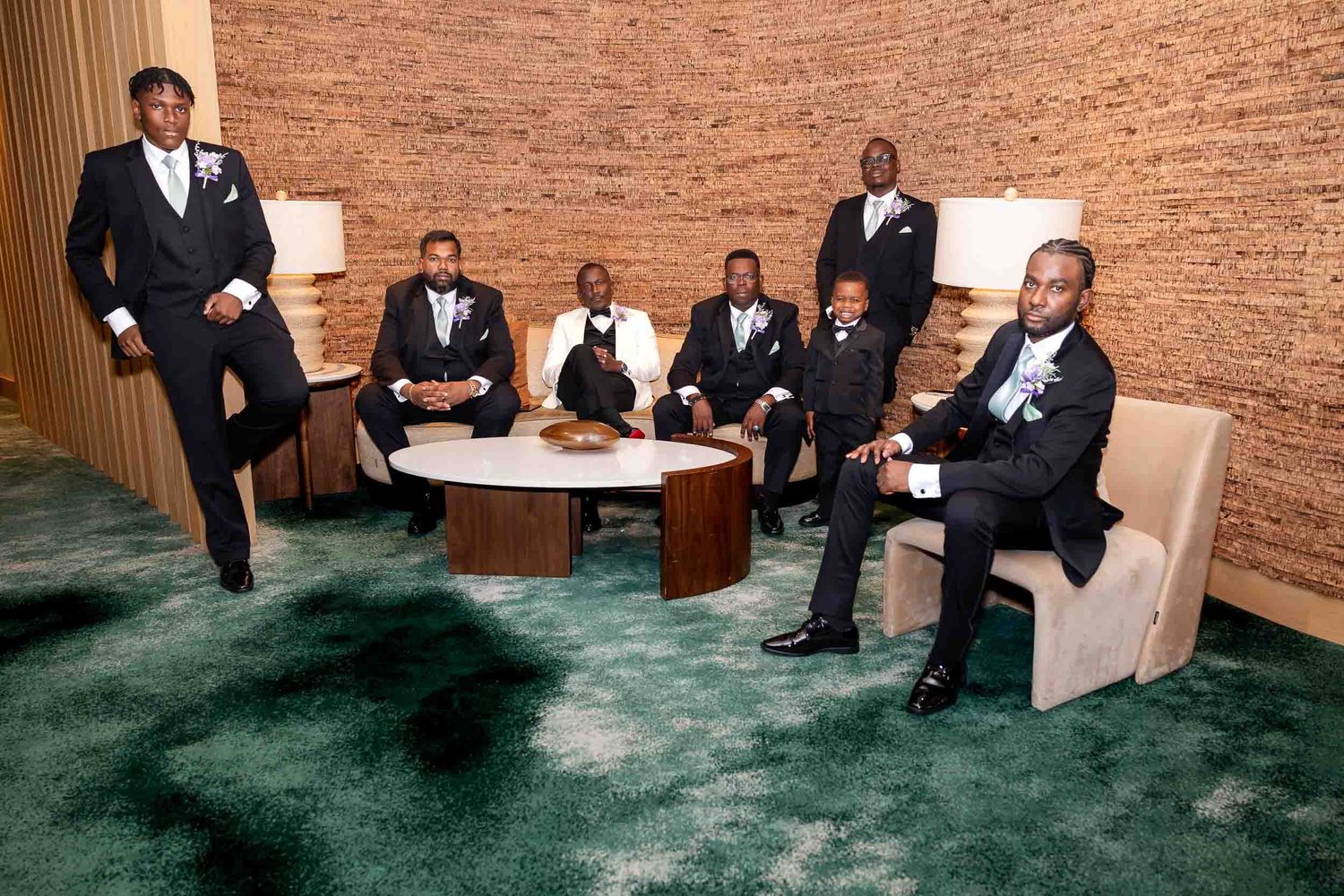 Seven well-dressed groomsmen in black tuxedos posing confidently in a modern lounge with green carpet.
