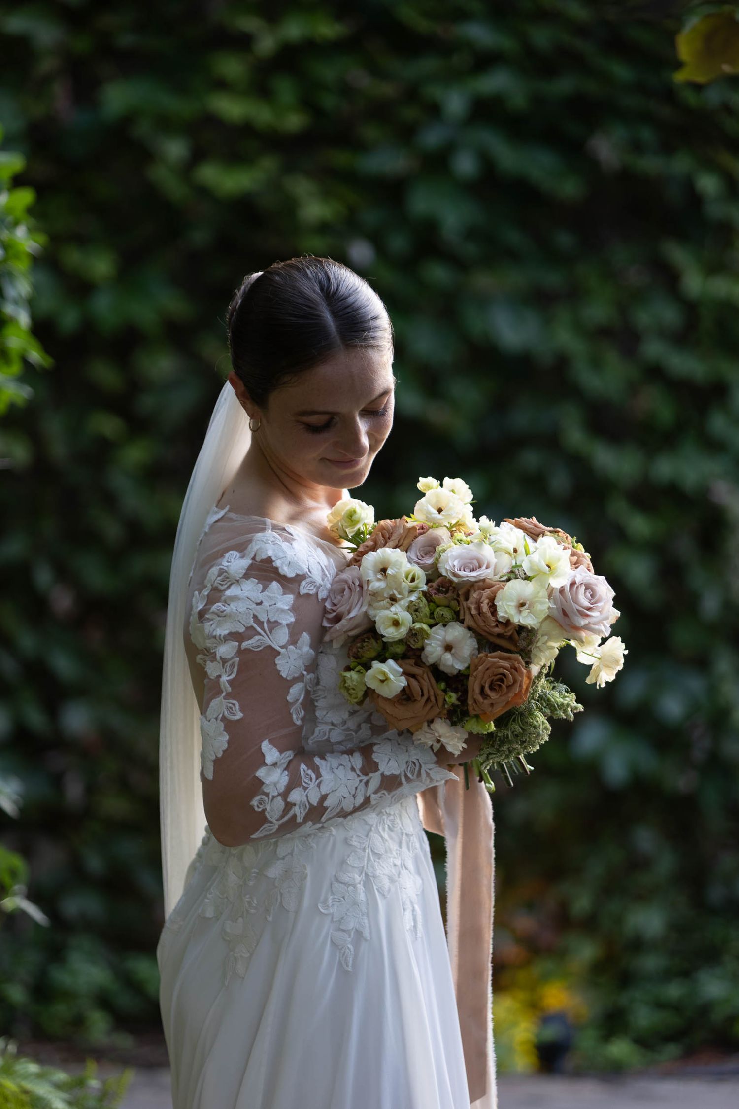 Bride in lace wedding dress holding bouquet of roses, standing before green ivy backdrop.