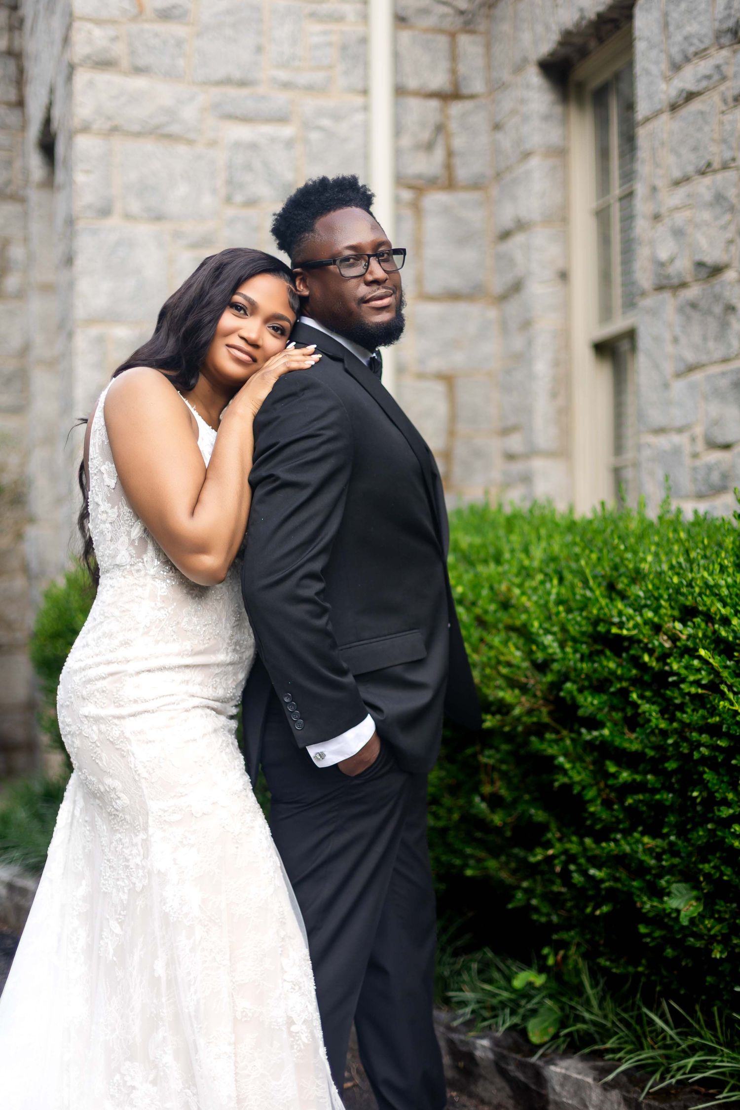 A couple poses together on their wedding day against a stone wall, wearing an elegant gown and black tuxedo.