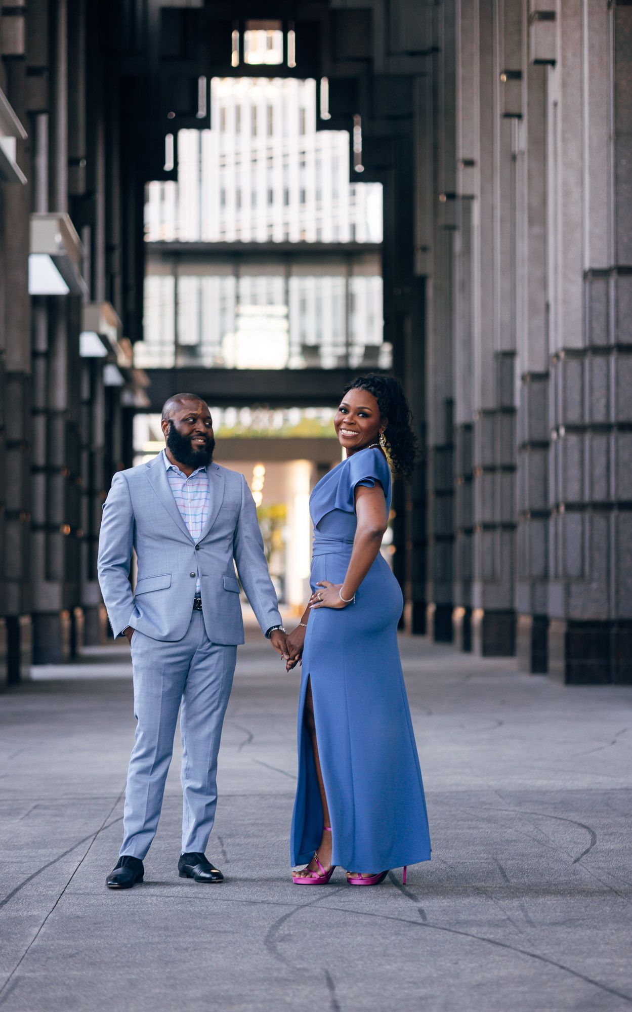 Smiling couple holding hands in a light blue suit and a blue slit dress with pink heels, posing between tall modern columns.