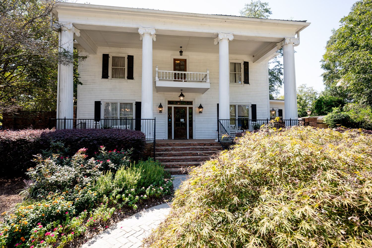 White two-story colonial home with large columns, black shutters, brick steps, and lush landscaped gardens.