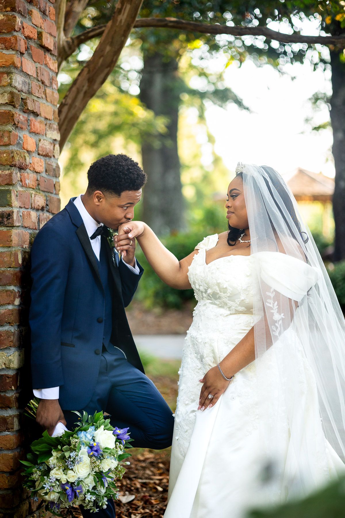 Groom in navy suit kisses bride's hand while she smiles in white lace gown and veil, holding blue floral bouquet outdoors.