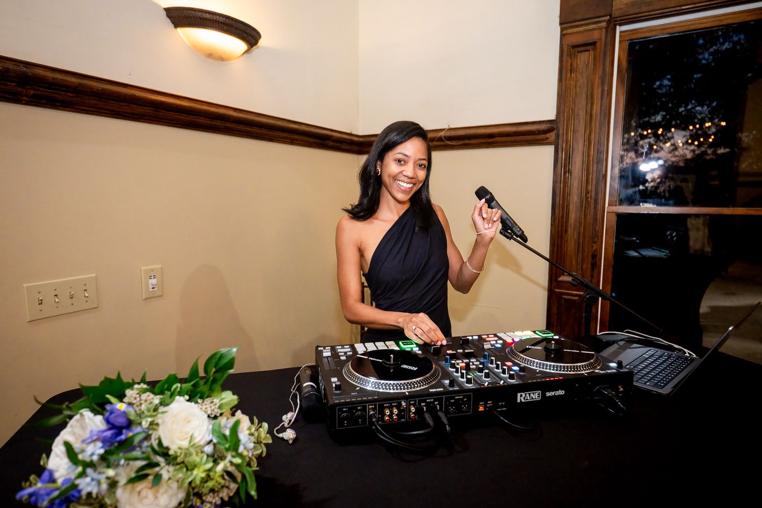 Female DJ in black dress smiling behind turntables holding microphone at elegant wedding reception.