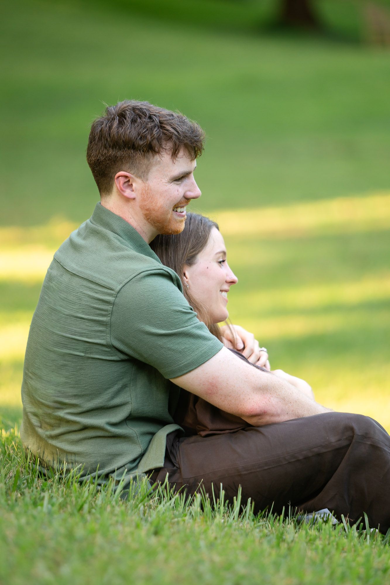 A smiling couple sits on green grass in a park, the man embracing the woman from behind in a warm, loving moment.