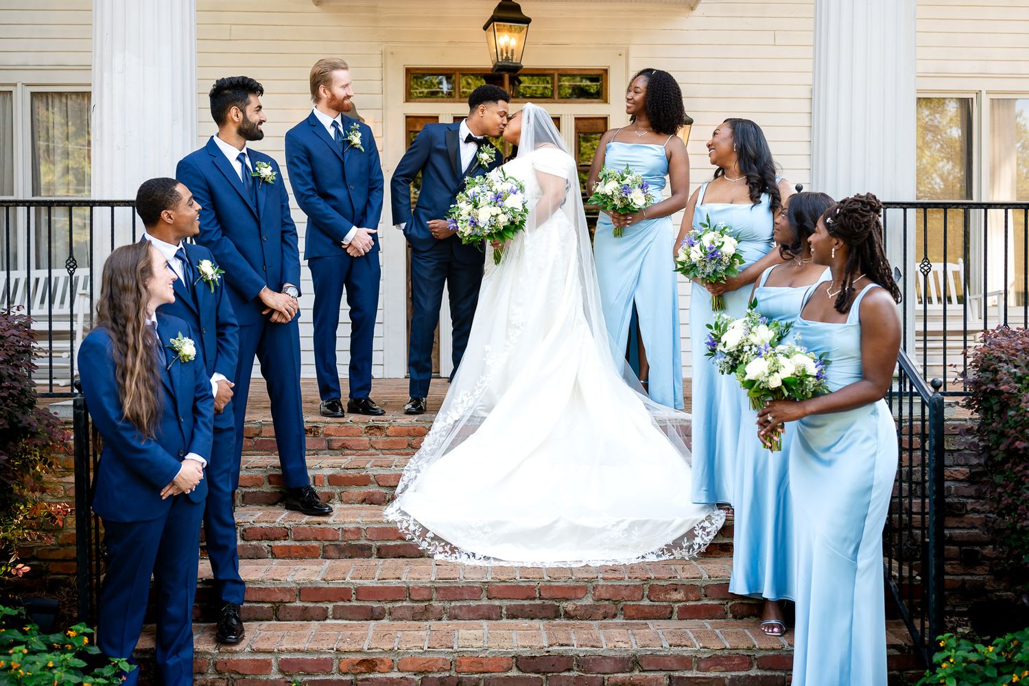 Wedding party poses on brick steps, bride in white gown with bridesmaids in blue dresses and groomsmen in navy suits.