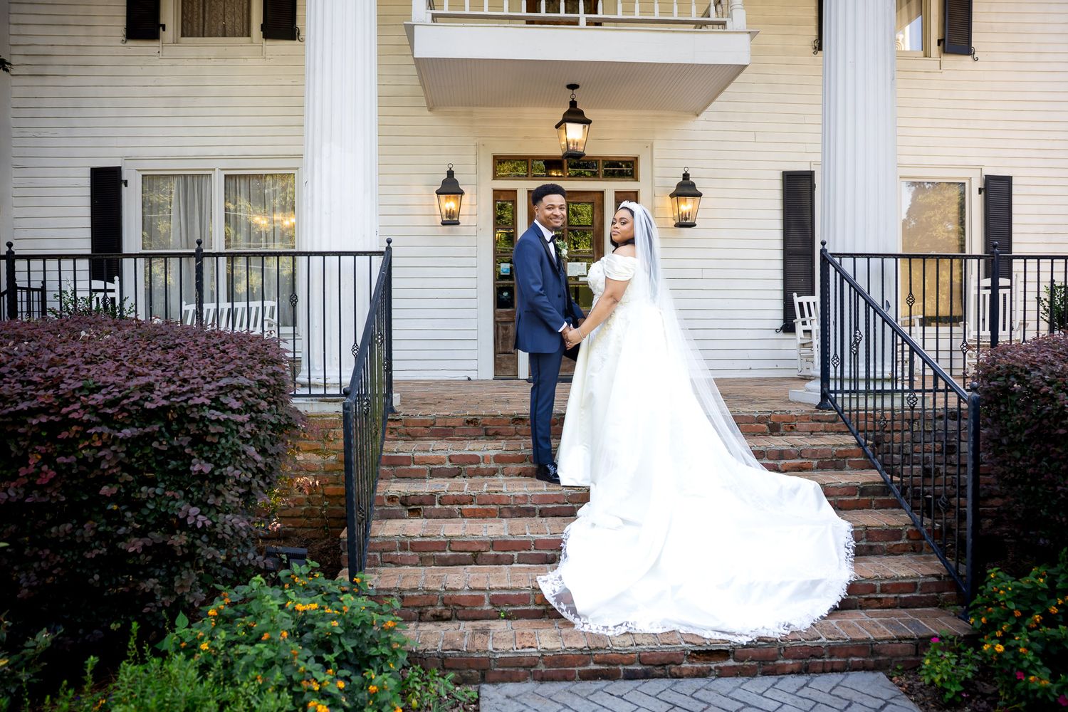Bride and groom holding hands on brick steps of elegant white colonial venue with black iron railings.