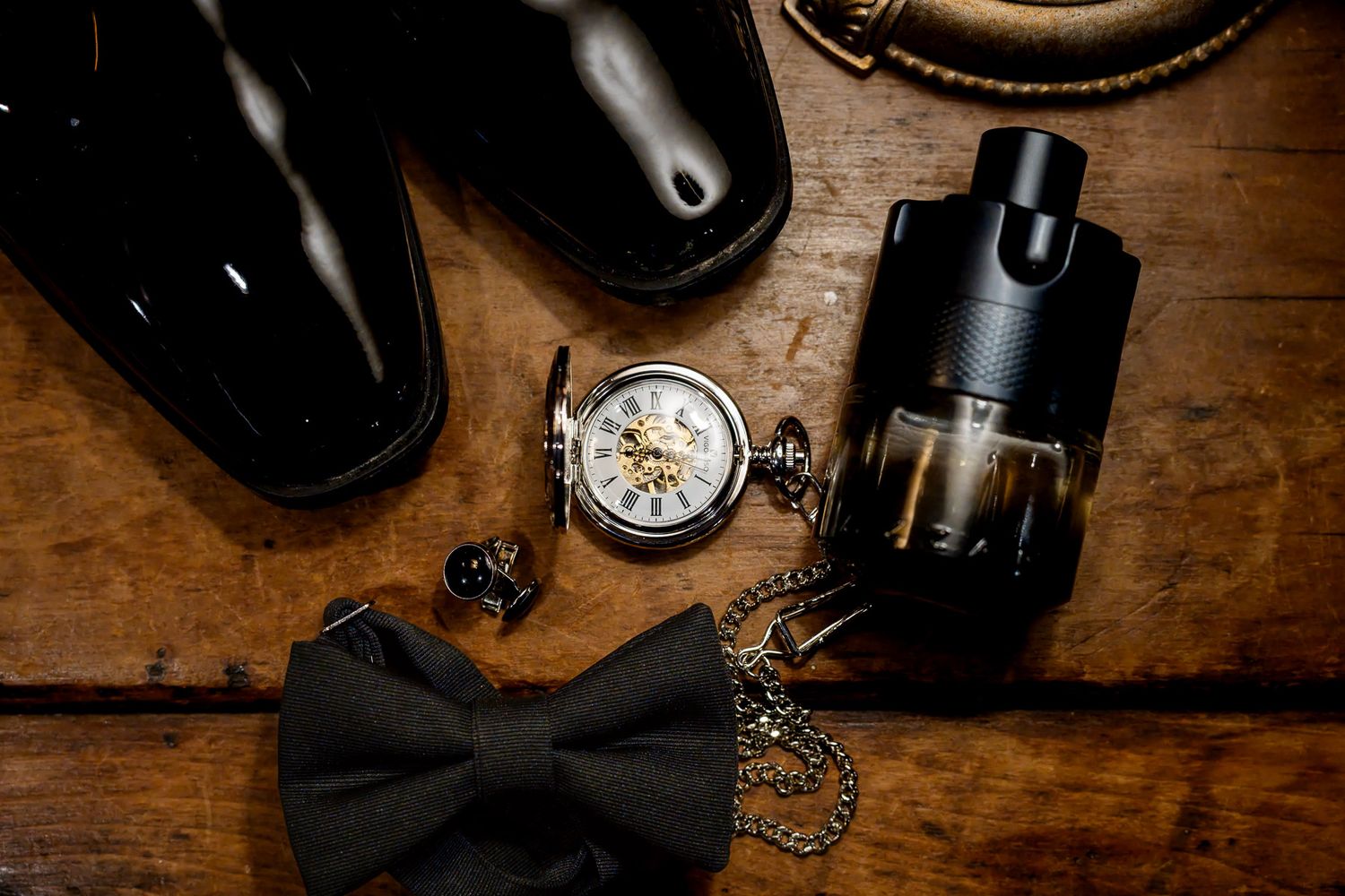 Elegant groom accessories flat lay featuring black patent shoes, pocket watch, bow tie, and cologne on rustic wood.