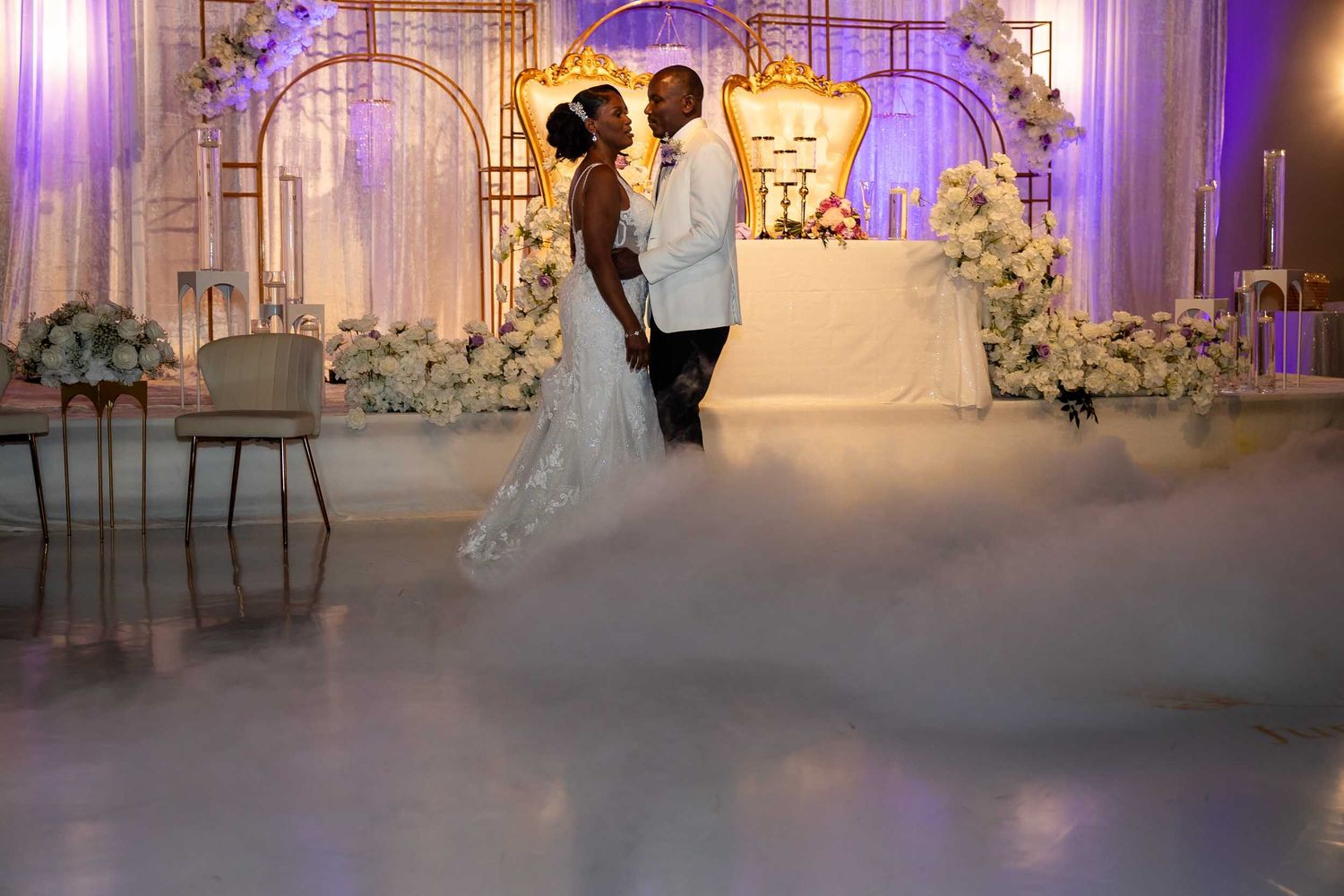 Bride and groom share first dance surrounded by dramatic fog on golden-accented wedding stage.