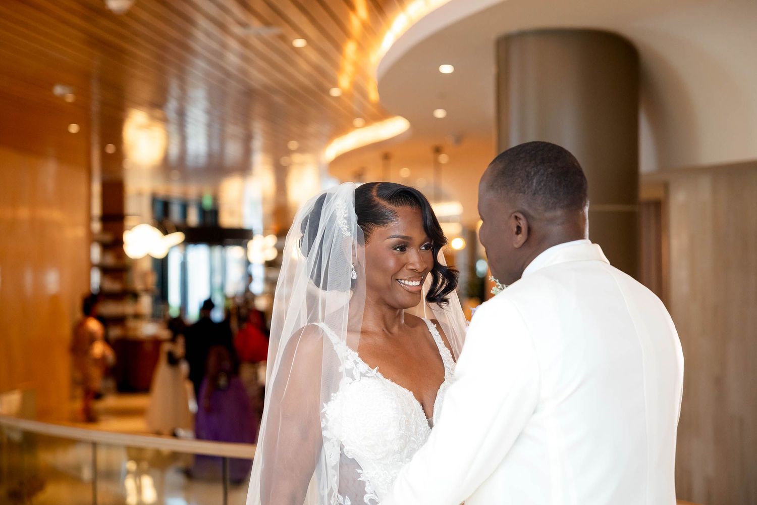 A Black bride in a white lace gown and veil smiles at her groom in a white suit in an elegant venue.