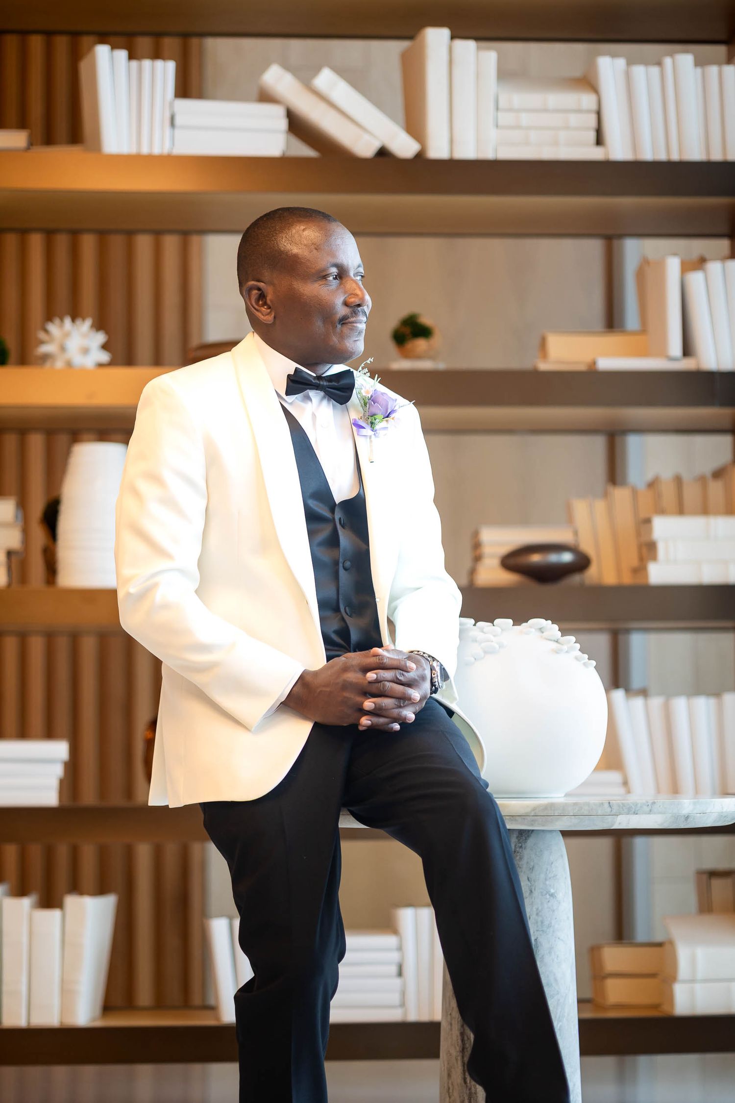 Elegant groom in white tuxedo jacket and black bow tie sitting by a bookshelf with a floral boutonniere.
