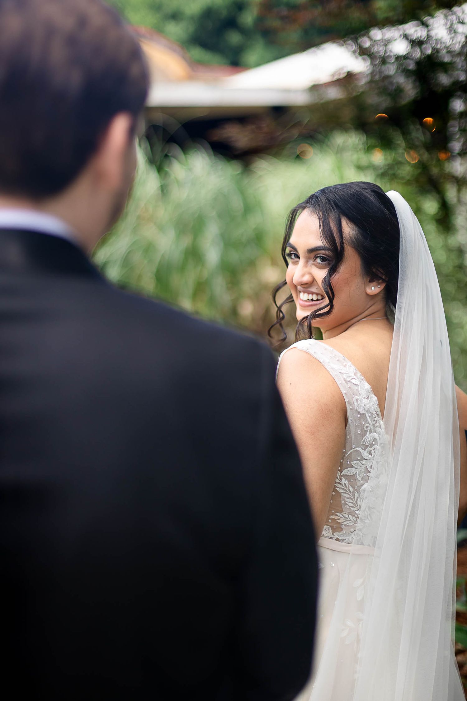 Smiling bride in white lace gown and veil glances back at groom during outdoor wedding ceremony.