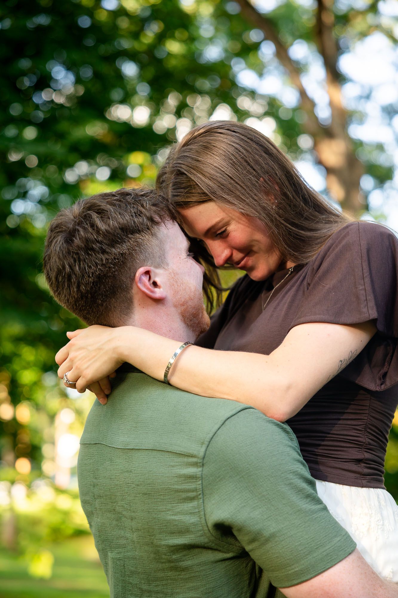 A couple embraces outdoors in a park, smiling at each other surrounded by lush green trees.