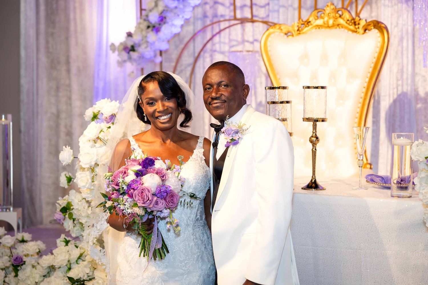 Smiling bride and groom posing at their wedding reception with white floral decor and gold throne chairs.