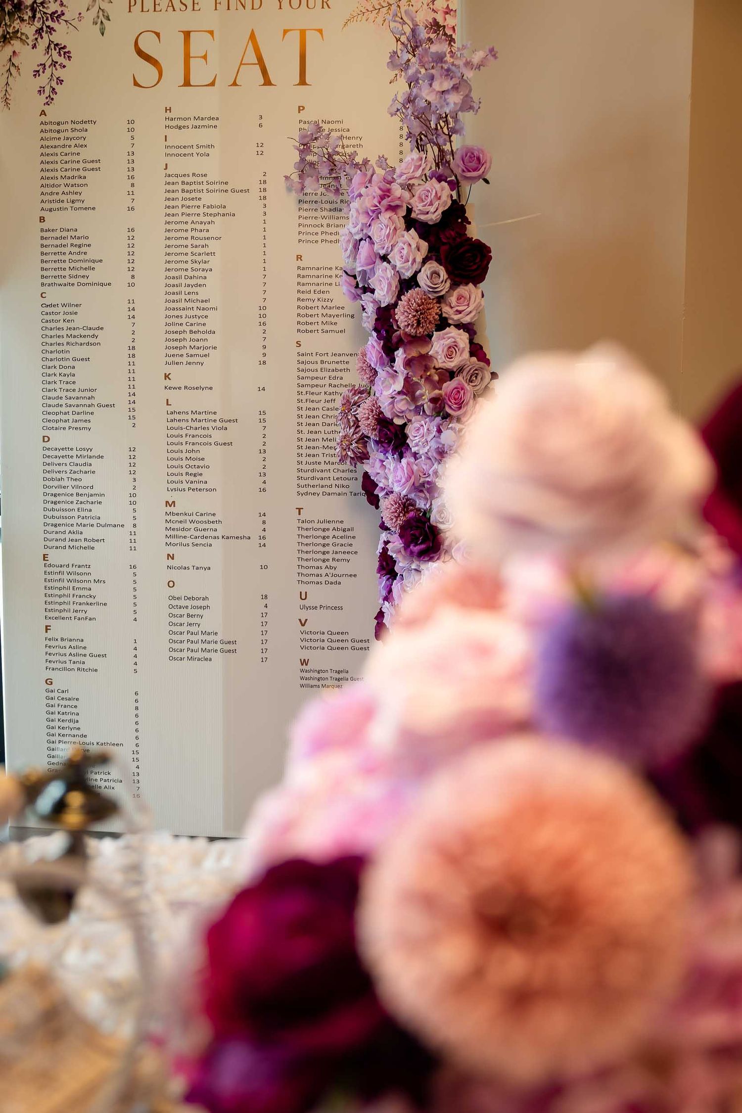 Floral-adorned wedding seating chart display with pink and purple blooms in the foreground at an elegant reception.