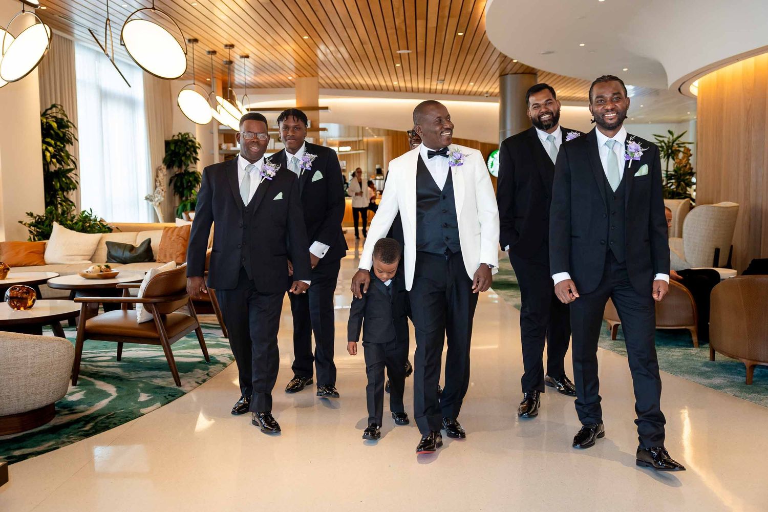 Groomsmen in black tuxedos and a groom in white vest walk with a young boy in a modern hotel lobby.