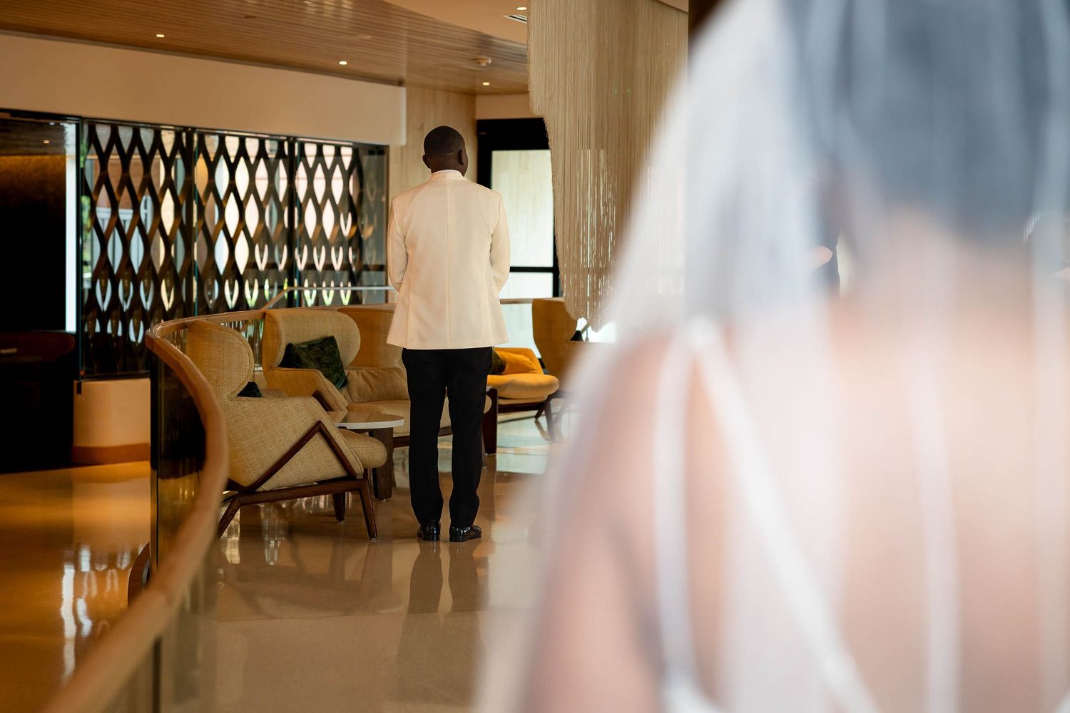 Bride in white veil approaches groom in white suit during romantic first look moment in elegant hotel lobby.