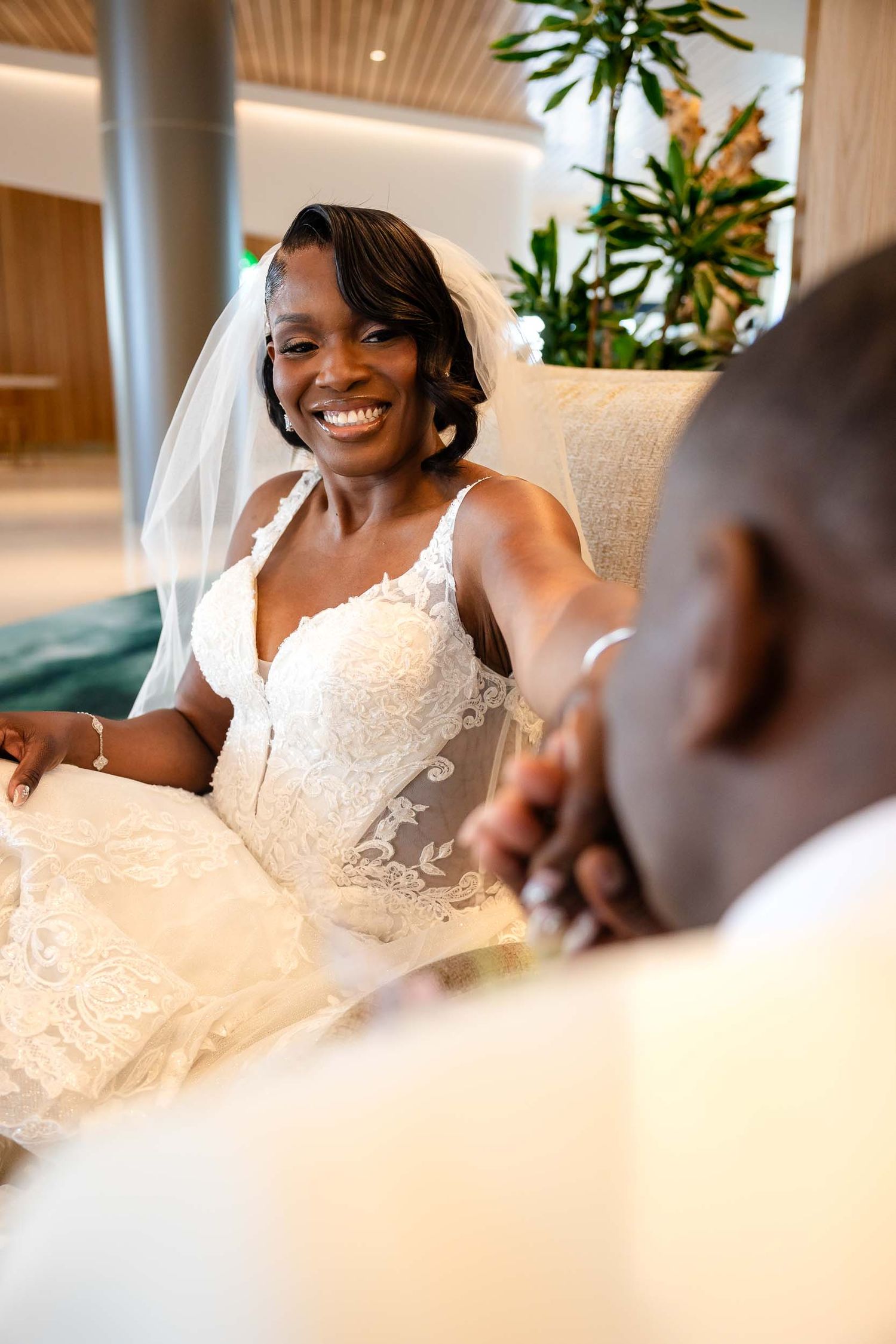 Radiant bride in lace wedding gown and veil smiles joyfully while holding groom's hand indoors.