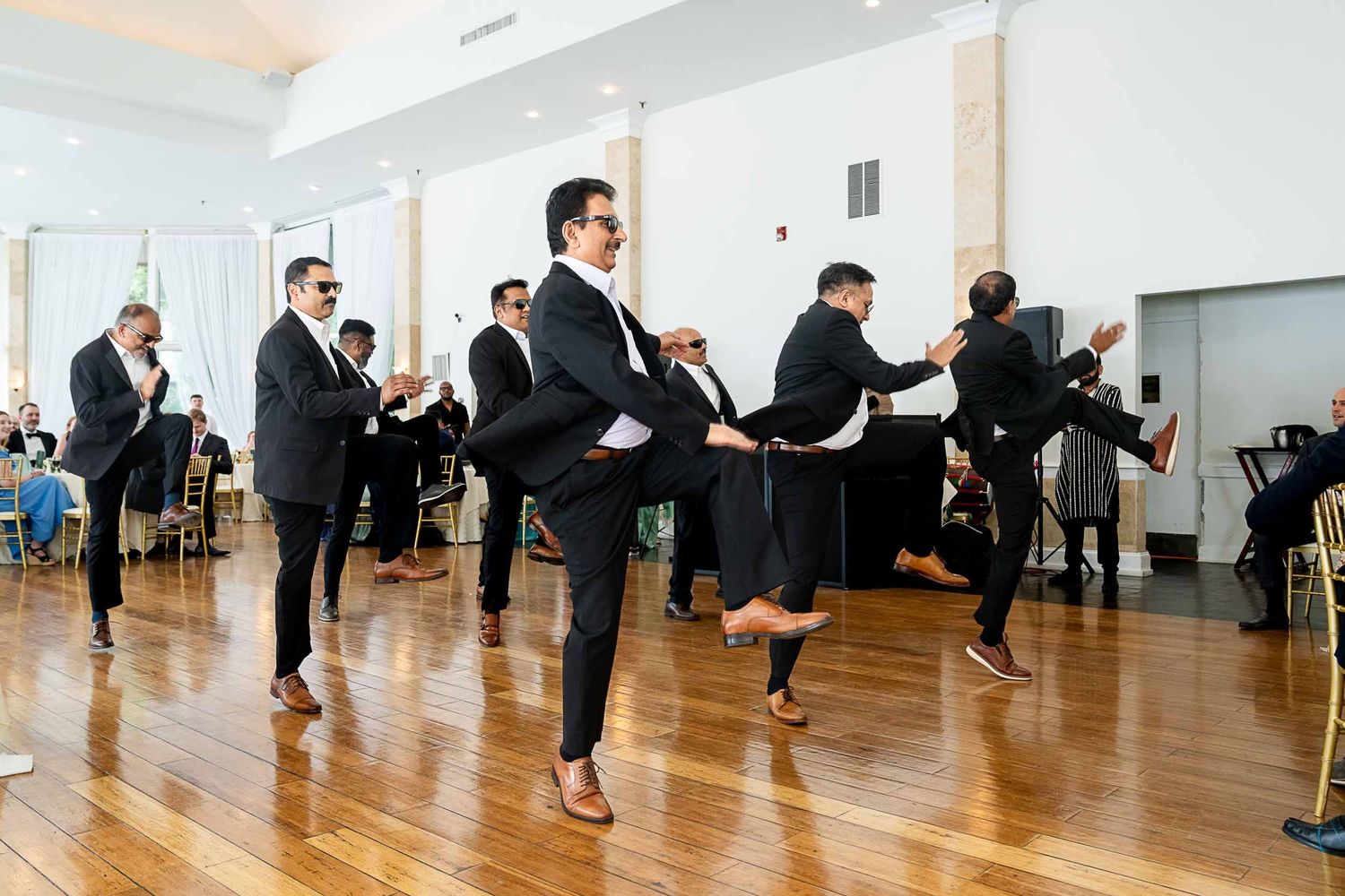 Groomsmen in black suits perform a lively traditional dance on a wooden dance floor at a wedding reception.