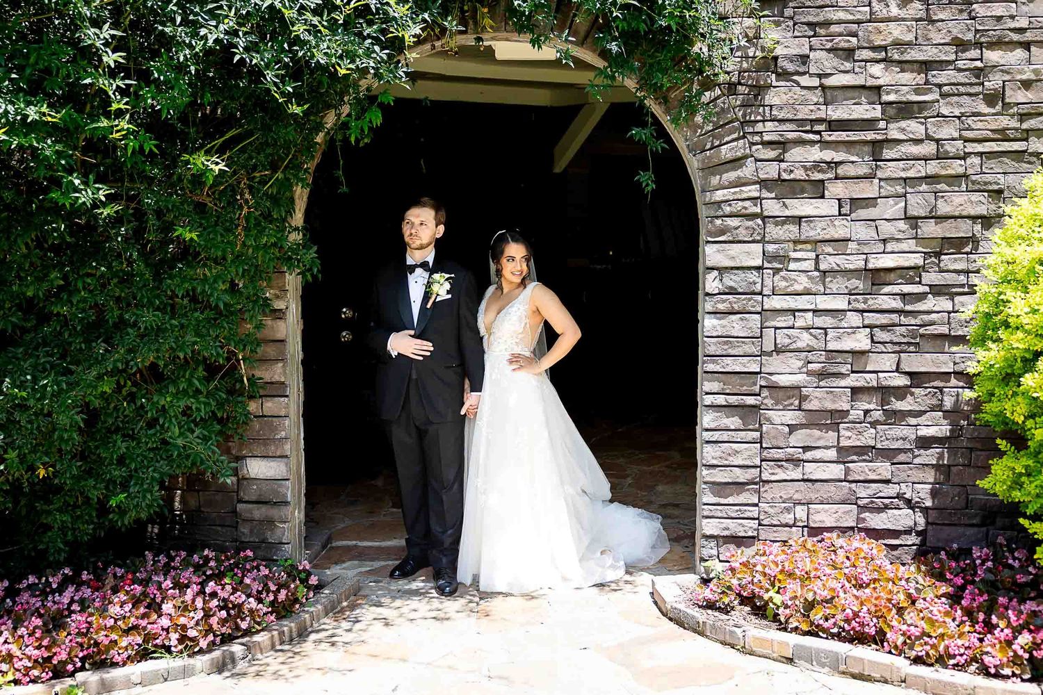 Bride and groom stand elegantly under stone arch surrounded by lush greenery and colorful flowers.