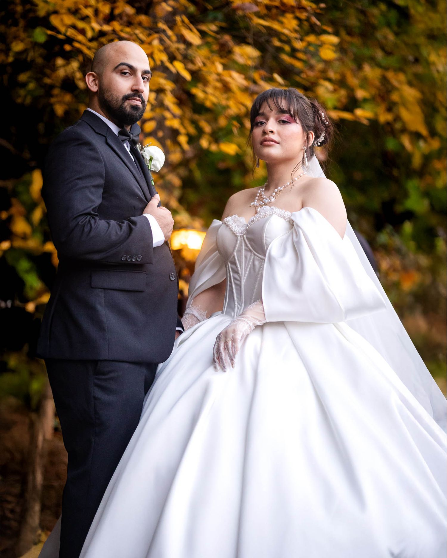 Bride in white ball gown and groom in black suit pose outdoors among autumn foliage for wedding portrait.