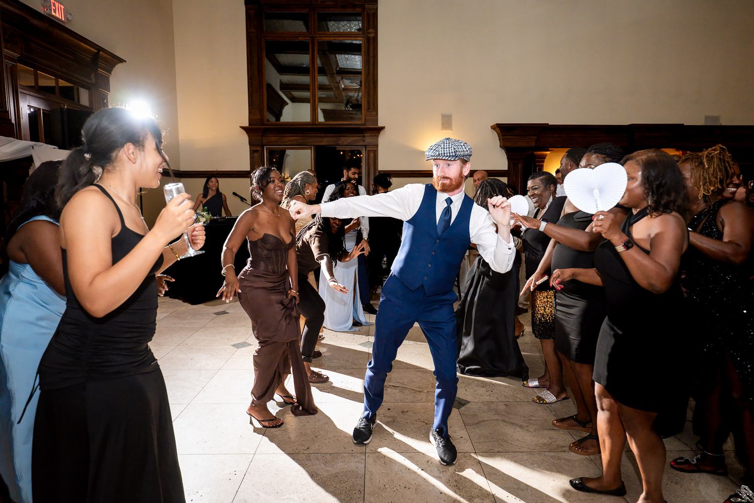 A man in a blue suit dances energetically at a wedding reception while guests cheer and wave fans around him.