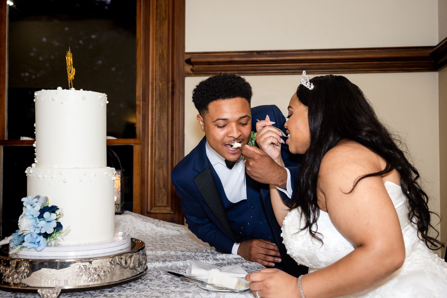 Bride and groom feeding each other wedding cake at reception table with white tiered cake and blue flowers.