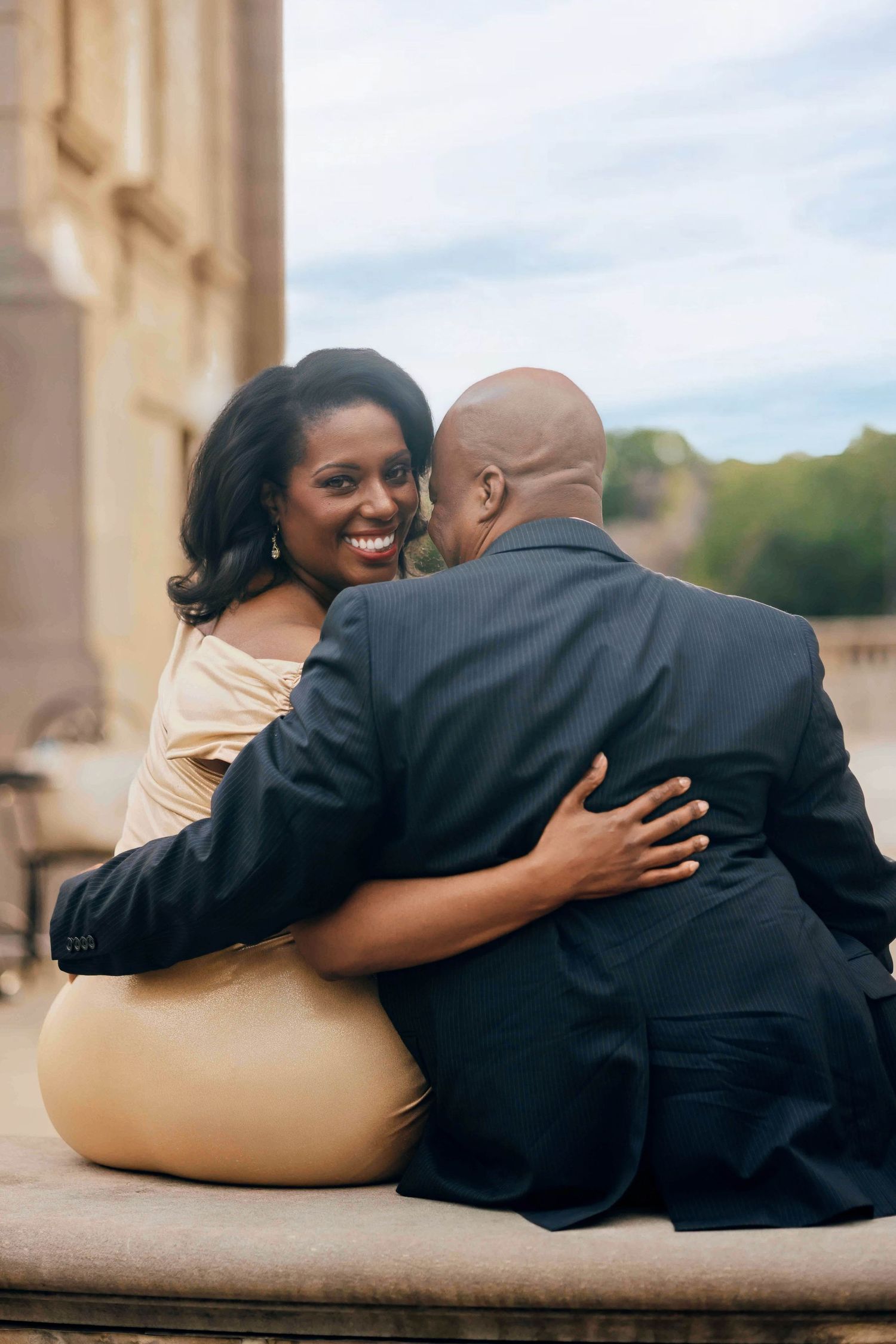 A smiling Black woman in a gold dress embraces a man in a dark suit while sitting outdoors at a elegant venue.