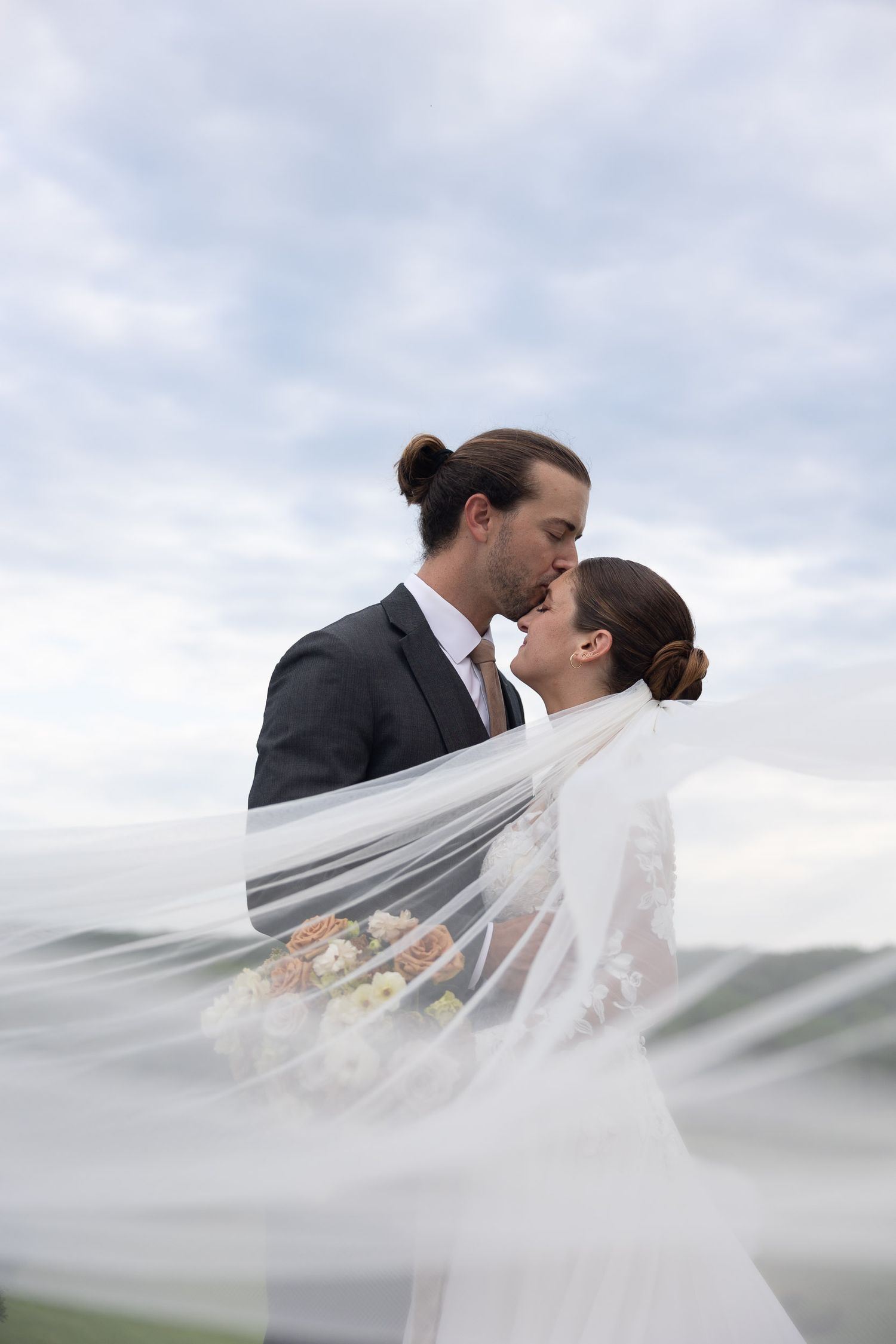 Bride and groom sharing a romantic kiss outdoors, with wedding veil billowing in the wind against a cloudy sky.