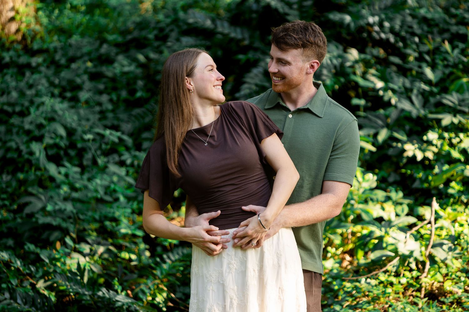 Couple smiling and holding hands outdoors surrounded by lush green foliage in a romantic engagement photo.