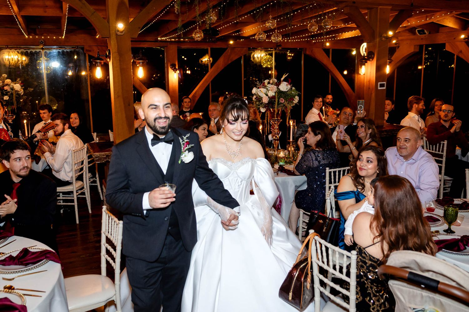 Bride and groom make grand entrance at elegant rustic wedding reception hall filled with guests.