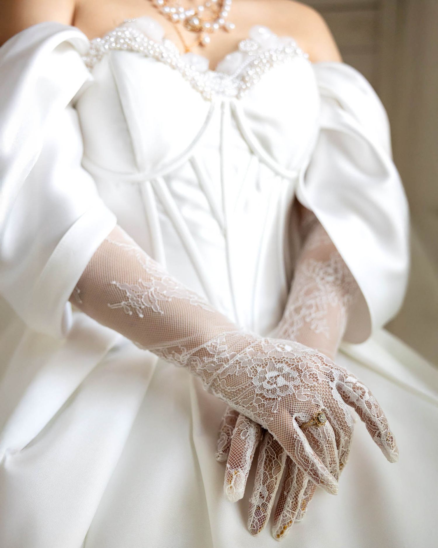 Bride wearing an off-shoulder white satin wedding gown with elegant lace gloves and pearl necklace.