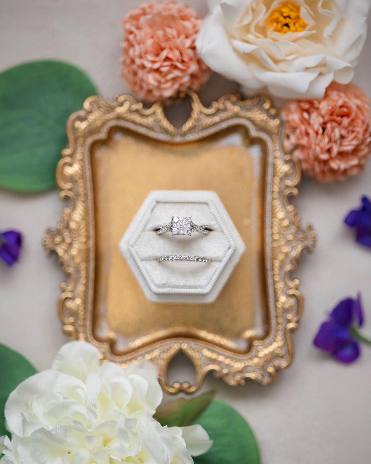 A diamond engagement ring displayed in a white hexagonal velvet box on a gold ornate frame surrounded by colorful flowers.