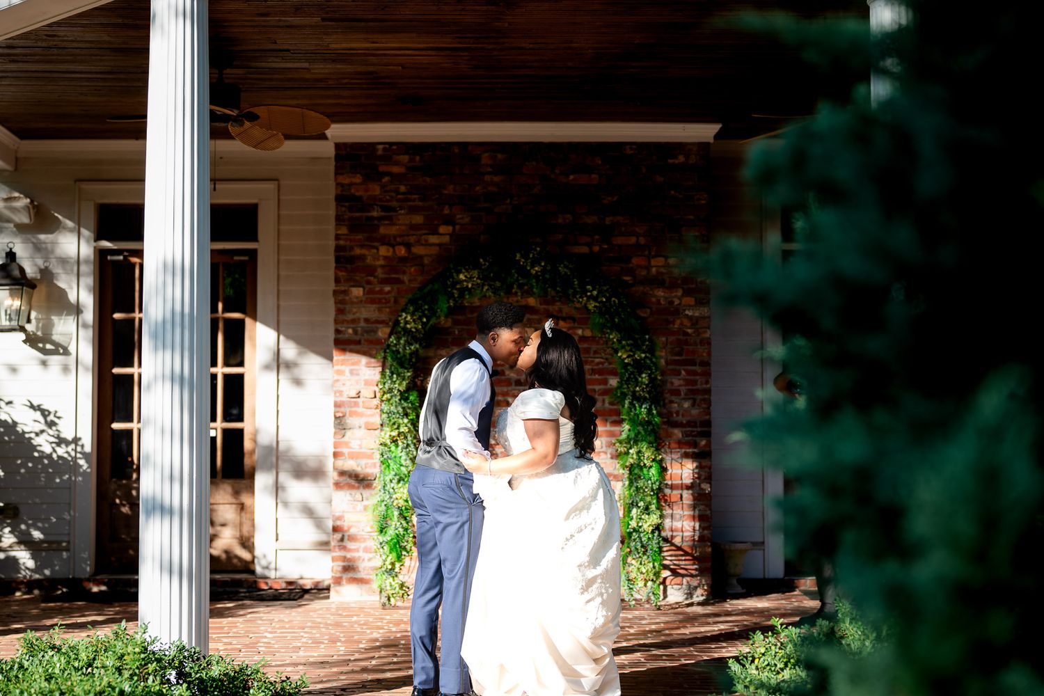 Bride and groom share a romantic kiss on a sunlit brick porch adorned with lush greenery at their wedding venue.