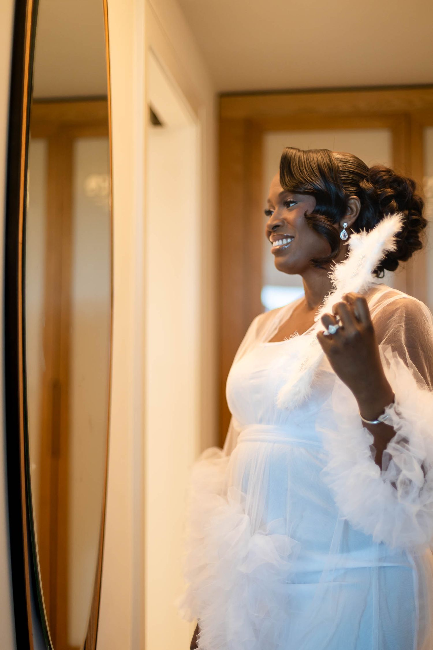 Radiant bride in white feather-trimmed wedding gown smiling at mirror reflection during bridal prep.