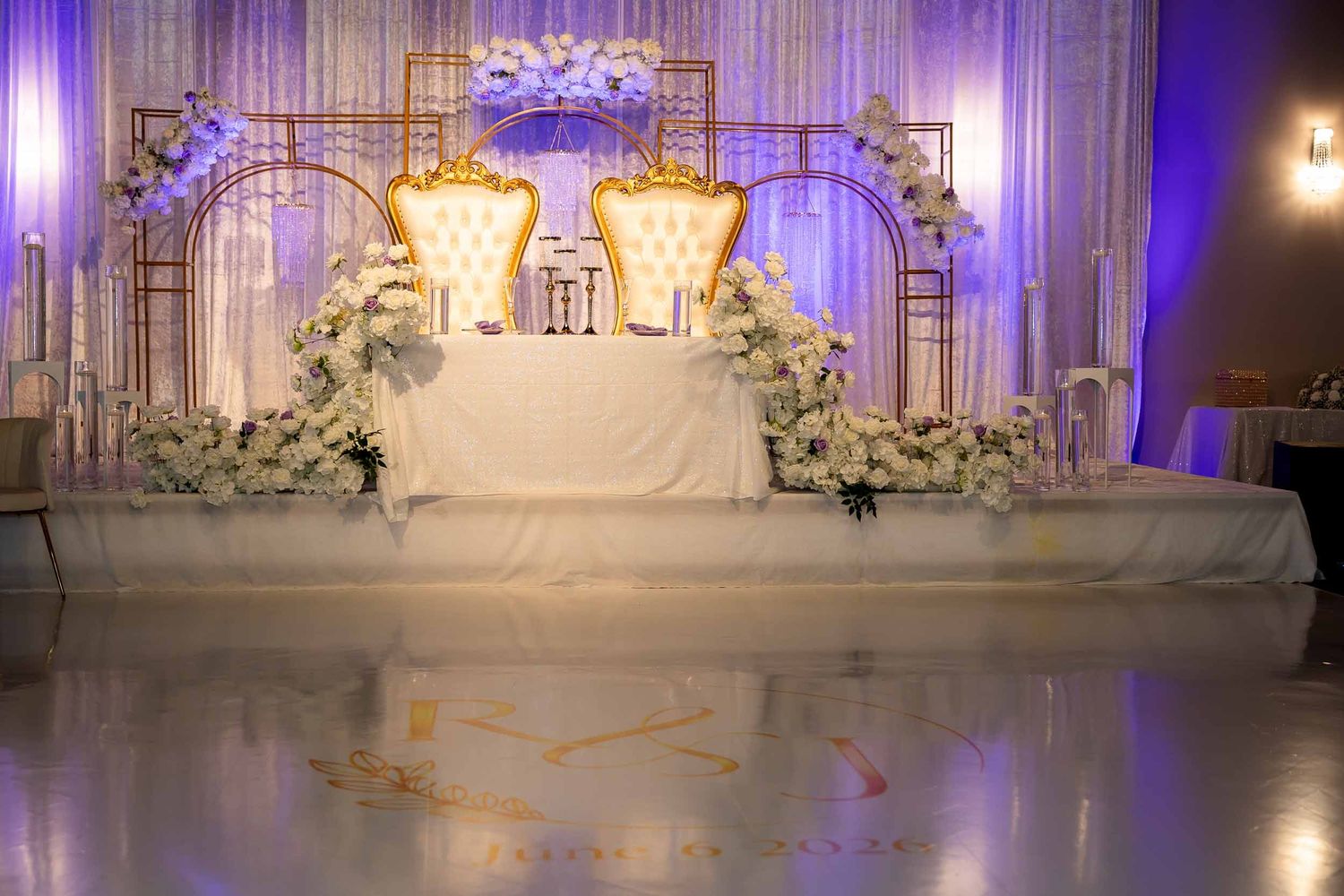 Elegant wedding sweetheart table with gold arch, white floral arrangements, and two ornate throne chairs on a shiny dance floor.