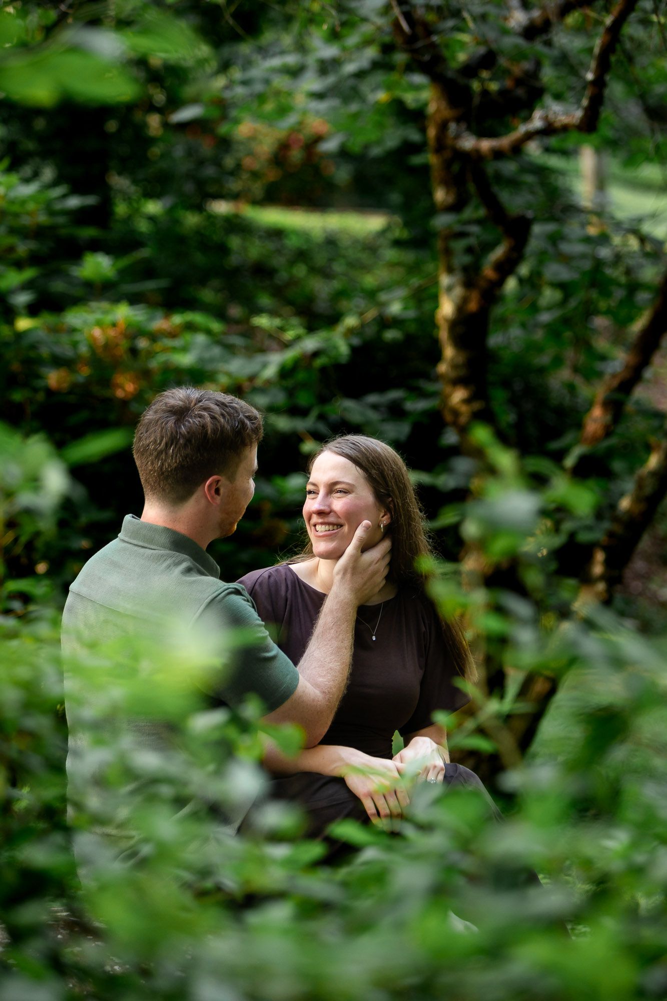 A couple shares a tender moment surrounded by lush green foliage, as a man gently touches a smiling woman's face.