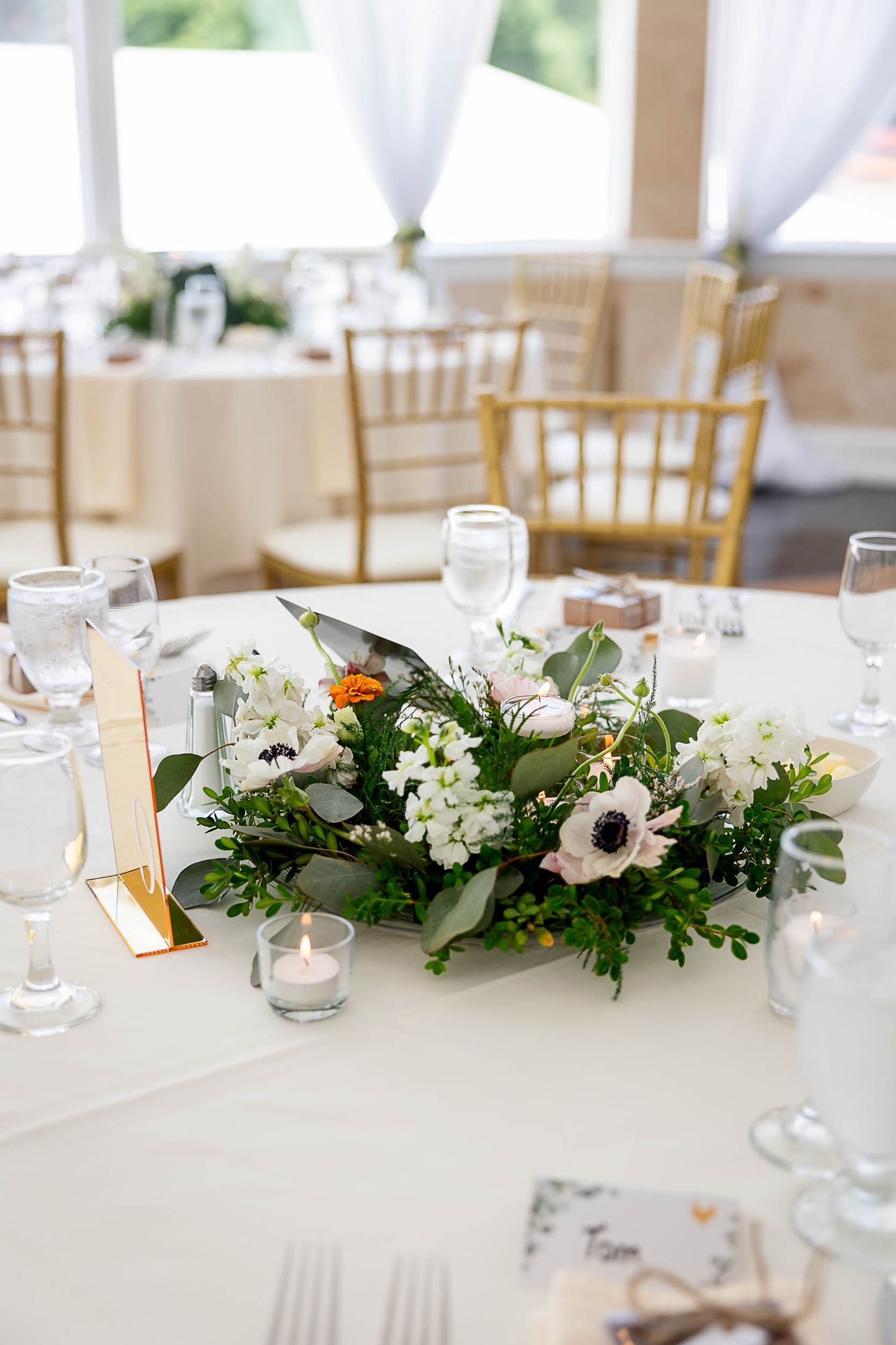 Elegant wedding reception table with white floral centerpiece featuring anemones and greenery, gold chairs, and glassware.