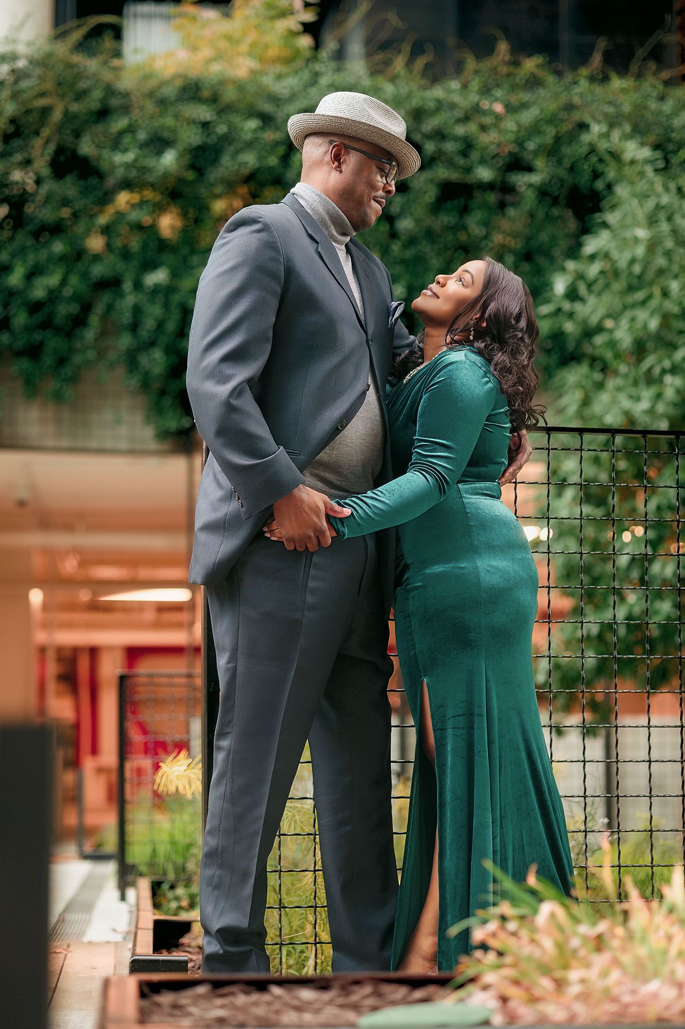 Couple in elegant attire, man in gray suit and hat, woman in green velvet gown, sharing a romantic moment outdoors.
